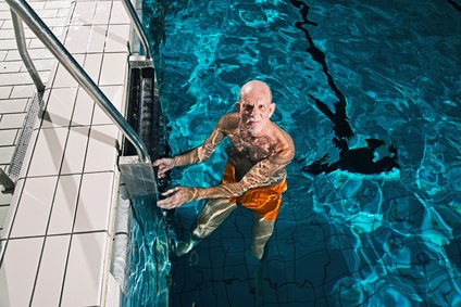 Healthy active senior man with beard in indoor swimming pool. Wearing orange swimming trunks.